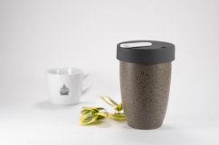 Loveramics Nomad Granit Thermotasse 250ml in gefleckter Sandfarbe mit dunkelgrauem Deckel, neben weißer Porzellanmasse mit Untertasse