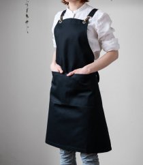 Delantal de barista negro con tirantes ajustables y aros metálicos dorados, elegantemente combinado con camisa blanca y jeans