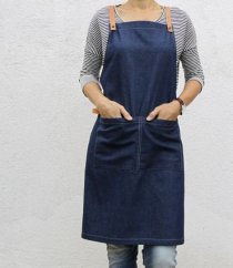Delantal de barista vaquero en azul oscuro con tirantes de cuero, mujer con camiseta a rayas con manos en bolsillos del delantal sobre fondo blanco