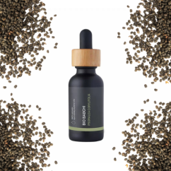 Tmavá skleněná lahvička s kapátkem a dřevěným uzávěrem BIO Babchi oleje obklopená rozptýlenými hnědými semínky na bílém pozadí