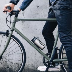 Mężczyzna w dżinsach trzymający zielony Kinto Travel Tumbler stalowy termiczny kubek na ramie miejskiego roweru, praktyczne podróżowanie z gorącymi napojami Mężczyzna w dżinsach trzymający zielony Kinto Travel Tumbler stalowy termiczny kubek na ramie miejskiego roweru, praktyczne podróżowanie z gorącymi napojami