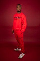 Muž v červené teplákové soupravě Tyson 2,0 s nápisem AMSTERDAM na mikině, stojí na červeném pozadí v bílých teniskách Adidas