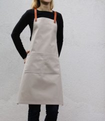 Detalle de delantal de barista color arena con tirantes de cuero y bolsillo práctico, modelo con camiseta negra y jeans