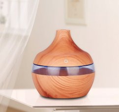 Aroma difuzér v dřevěném provedení s kulatým tvarem a průhledným pruhem uprostřed, umístěný na bílé polici v světlém interiéru Aroma difuzér v dřevěném provedení s kulatým tvarem a průhledným pruhem uprostřed, umístěný na bílé polici v světlém interiéru