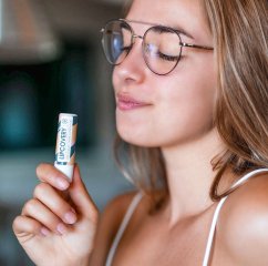 Žena s brýlemi drží v ruce bílou tubu Harmony Lipcovery CBD balzámu na rty s barevným vícebarevným obalem Žena s brýlemi drží v ruce bílou tubu Harmony Lipcovery CBD balzámu na rty s barevným vícebarevným obalem