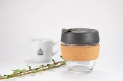 KeepCup Brew Cork Nitro S, cană ecologică din sticlă cu bandă de plută și capac gri de 227 ml, lângă o ceașcă albă de porțelan cu o ramură pe fundal