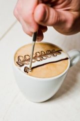 Ruka držící stříbrné Motta baristické pero při vytváření latte art vzoru na bílé pěně cappuccina v bílém šálku na dřevěném povrchu
