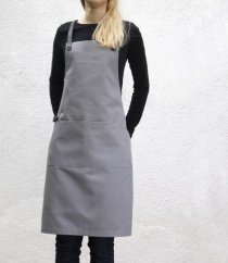 Delantal de barista gris con detalles de cuero sobre una mujer con camiseta negra, diseño elegante con bolsillo y correas ajustables para cocina y café