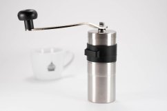 Porlex Mini II mlýnek na kávu z nerezové oceli s černým silikonovým pásem, vedle bílého hrnku s logem a dlouhou kovovou rukojetí