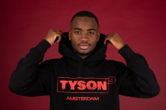 Černá mikina Tyson 2,0 vel. XL s kapucí a červeným nápisem TYSON AMSTERDAM, model pózuje s rukama u kapuce na červeném pozadí