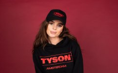 Černá kšiltovka Tyson 2,0 s červeným logem TYSON AMSTERDAM na čelním panelu, nosí ji žena v černé mikině na červeném pozadí