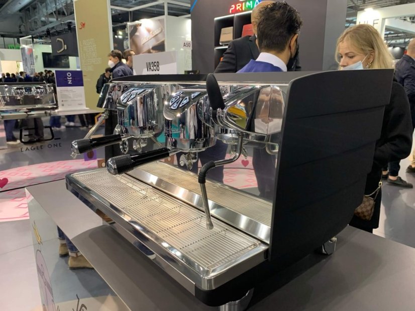 Kávovar Victoria Arduino White Eagle na výstavě s návštěvníky v pozadí, ukazující praktické použití stroje v profesionálním prostředí