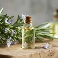 Sticlă mică de sticlă cu dop de plută, umplută cu ulei transparent, poziționată pe o placă de lemn lângă flori mov și rozmarin verde